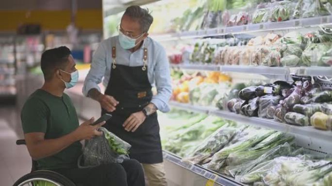周末，坐在轮椅上的亚洲印度残疾男子在超市的智能手机上向高级男子零售助理询问购物清单