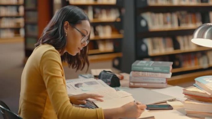 一名学生在校园图书馆学习考试或研究项目。一个快乐的年轻律师，戴眼镜坐在办公桌前，拿着一堆书，一边读教