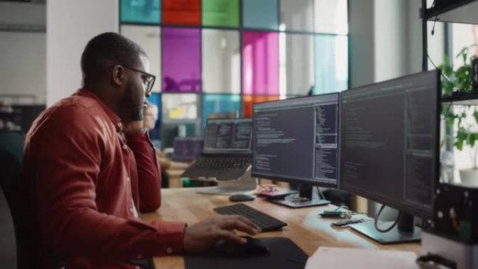 黑人在台式电脑上写代码行，电脑上有两台显示器和一台笔记本电脑，放在时尚的办公室里。专业男性开发人员为