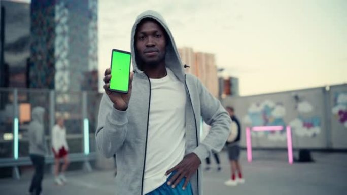 英俊的黑人拿着智能手机，显示绿屏模拟显示。年轻时尚的非洲男子站在城市环境中，不同的人在背景中踢街头足