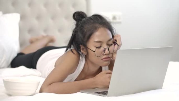 年轻漂亮的眼镜亚洲女人躺在白色卧室。女孩在使用笔记本电脑时小睡一会儿。检疫期间在家穿着便服的昏昏欲睡