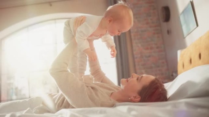 可爱的年轻美丽的母亲在床上玩耍和抚慰可爱的婴儿。微笑的有爱心的妈妈和可爱的小婴儿在卧室里拥抱。母亲温