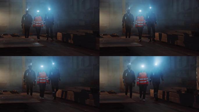 三名多元文化的重工业工程师和工人在安全帽上使用手电筒走在深色钢铁工厂。工业承包商戴着安全Unofir