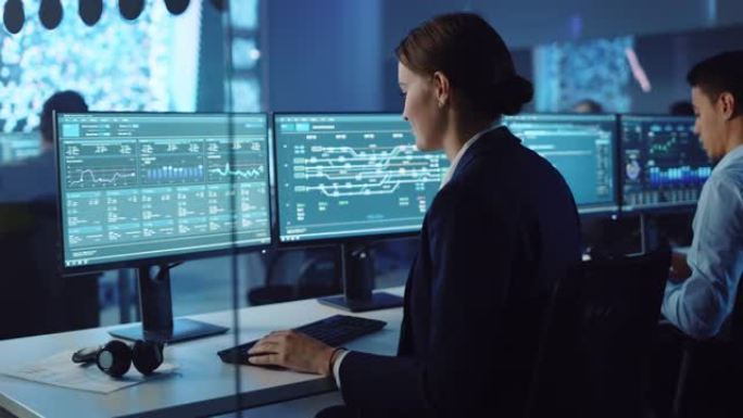 自信的女性数据科学家在神经网络的大型基础设施控制和监控室中的个人计算机上工作。女工程师和同事在办公室