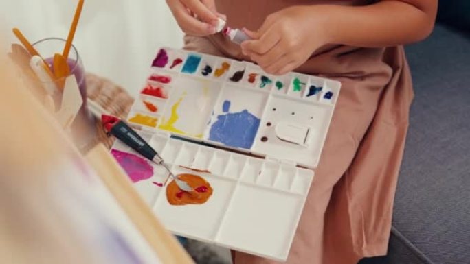 特写亚洲学步女孩围裙坐在油画画布前的沙发椅把丙烯酸色放在调色板上在家客厅使用调色板刀混合颜色。
