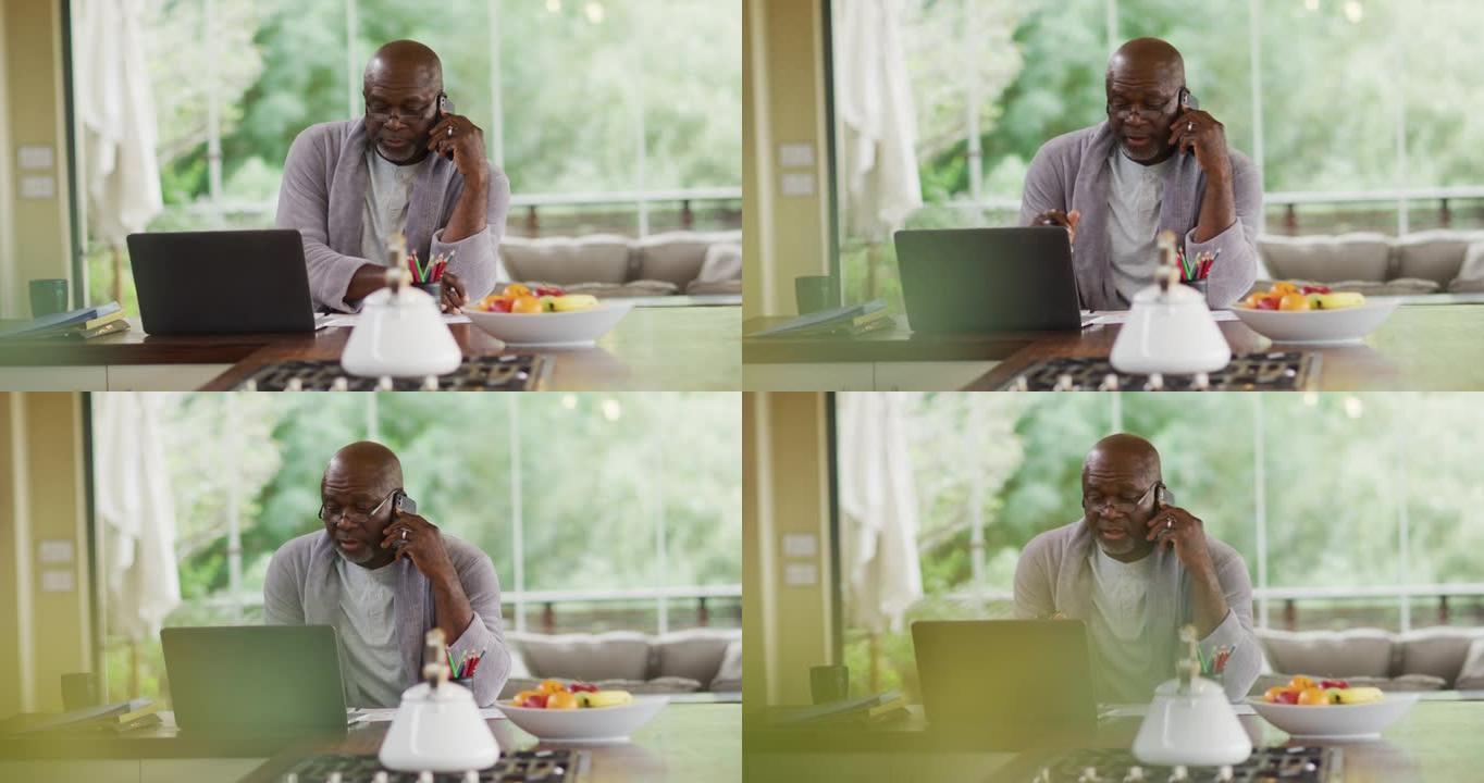 穿着浴袍的非洲裔美国高级男子坐在厨房里使用笔记本电脑并在智能手机上聊天
