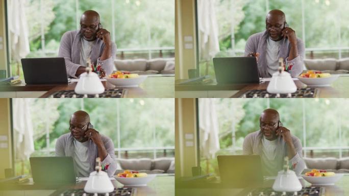 穿着浴袍的非洲裔美国高级男子坐在厨房里使用笔记本电脑并在智能手机上聊天