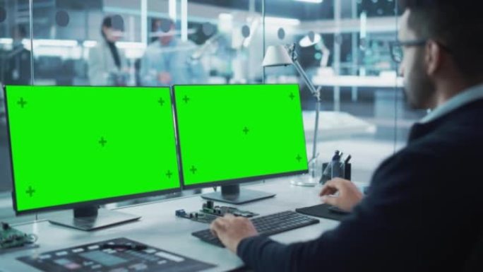 工业工程师在现代技术研发中心的台式计算机上工作，该计算机具有两个绿屏Chromakey显示屏。肩膀上