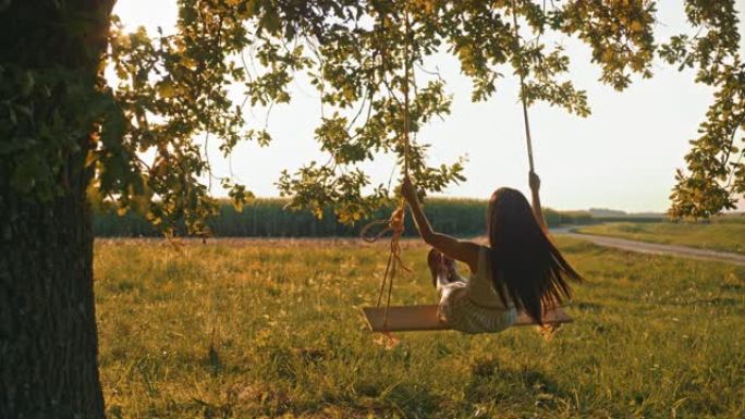 慢动作，手持镜头拍摄一个穿着白色连衣裙的年轻女孩摇摆，坐在悬挂在阳光充足的草地上的大树枝上的绳索秋千