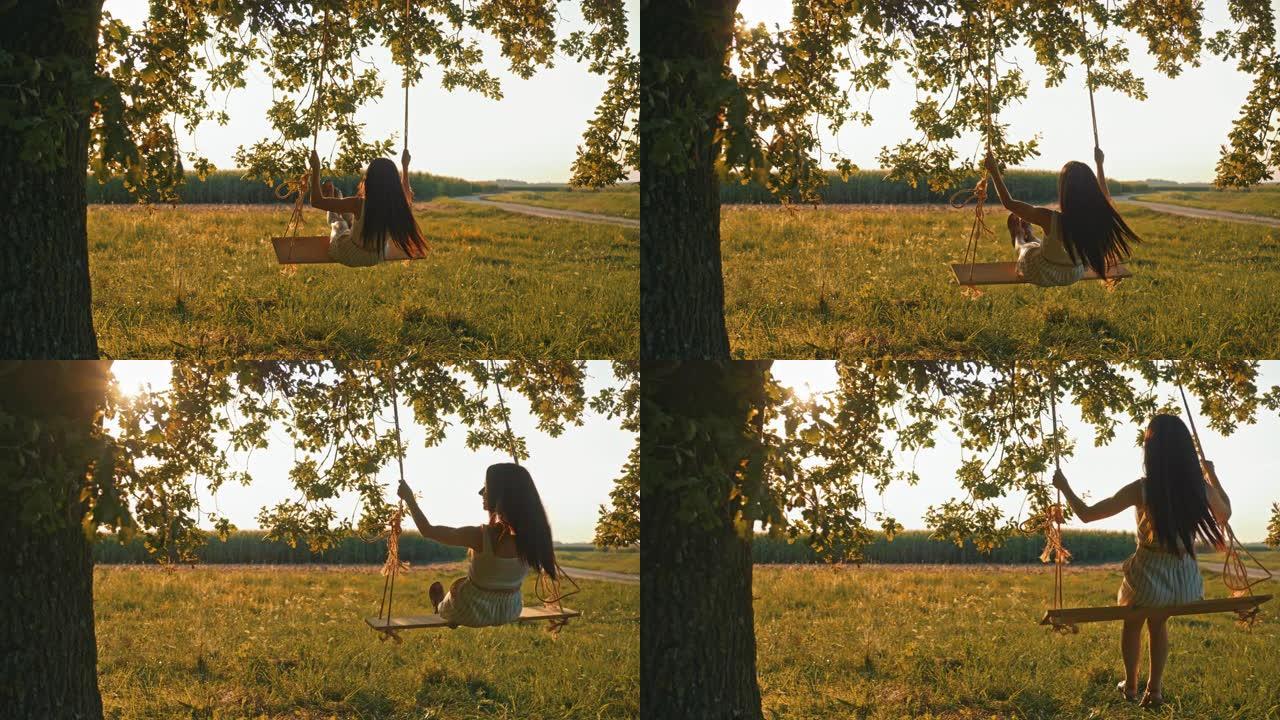 慢动作，手持镜头拍摄一个穿着白色连衣裙的年轻女孩摇摆，坐在悬挂在阳光充足的草地上的大树枝上的绳索秋千
