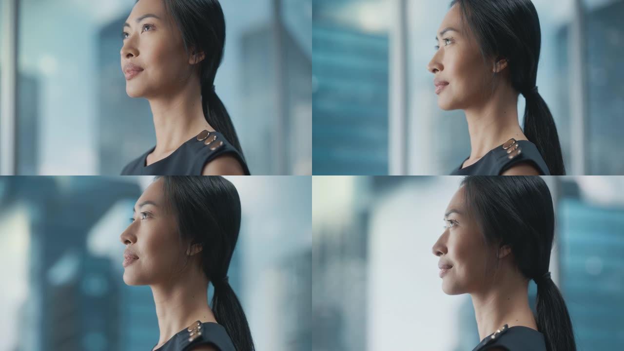 美丽的亚洲女商人肖像，穿着时髦的黑色连衣裙，在摩天大楼的大城市办公室的窗户旁边摆姿势。自信的女CEO