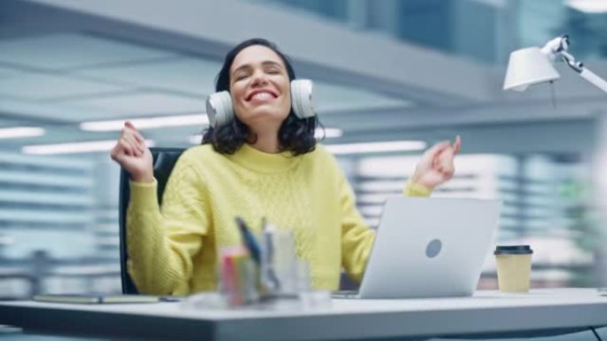 360学位办公室: 微笑的西班牙裔女商人坐在办公桌前在电脑上工作，戴着耳机，听音乐，跳舞。她很快乐，