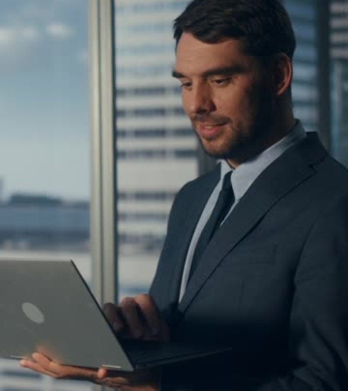 垂直屏幕: 商人正站在办公室，使用笔记本电脑，在具有大城市景观的窗户旁边。成功的公司首席执行官或财务