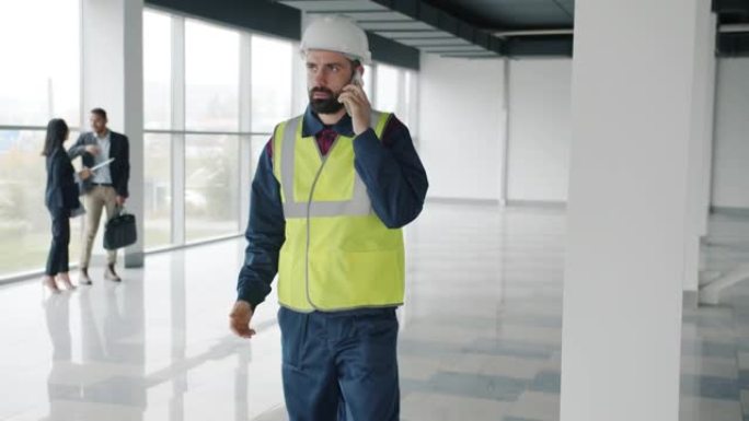 在新楼里，戴着头盔和背心的工程师在手机上讲话的慢动作肖像