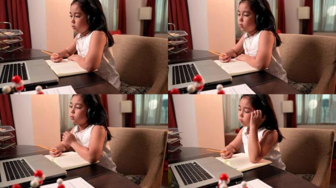 亚洲女孩在通过笔记本电脑和耳机进行在线学习时感到无聊。社交距离，儿童教育，自学，电子学习，家庭教育概