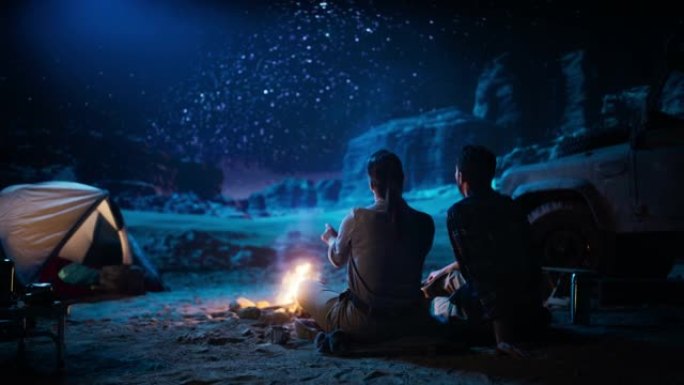 幸福的情侣大自然在峡谷中露营，坐在篝火旁看着夜空，银河中充满明亮的星星。两个恋爱中的旅行者在浪漫的假