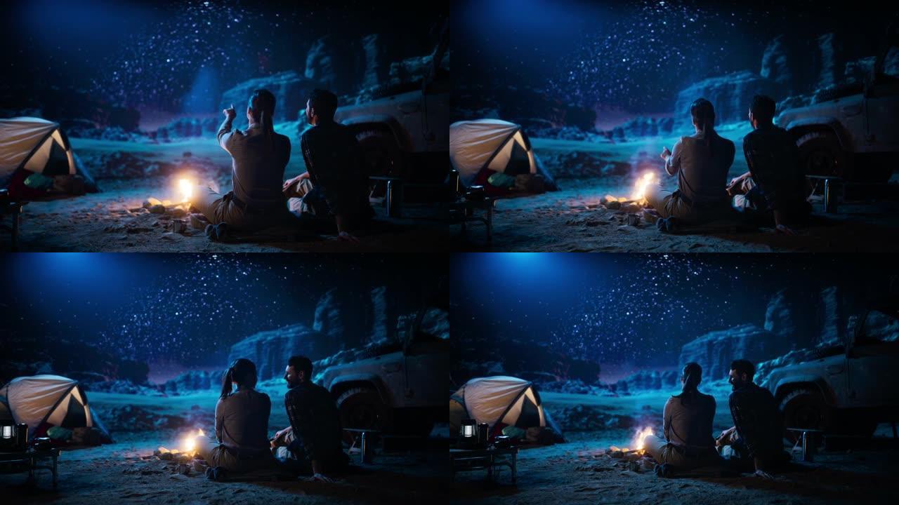 幸福的情侣大自然在峡谷中露营，坐在篝火旁看着夜空，银河中充满明亮的星星。两个恋爱中的旅行者在浪漫的假