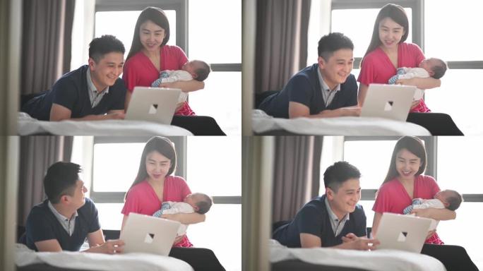 亚洲华裔父亲躺在床上使用笔记本电脑在线购物，而母亲则在卧室里抱着新生的男婴