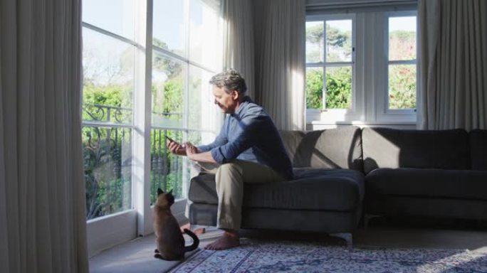 快乐的白人老人坐在阳光明媚的客厅里，用智能手机抚摸猫