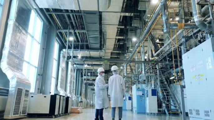 两名身穿实验室外套的专家正在工厂单位穿行。工厂设施，在现代工业工厂工作的工程师。