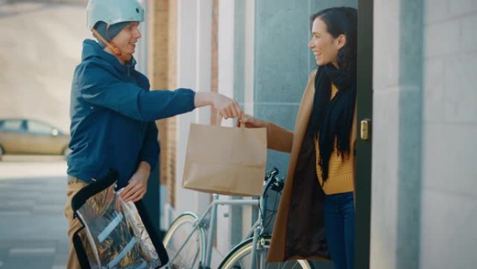 快乐的送餐男子在自行车上穿着保暖背包，向一位美丽的女性顾客提供餐厅订单。Courier在现代城市地区
