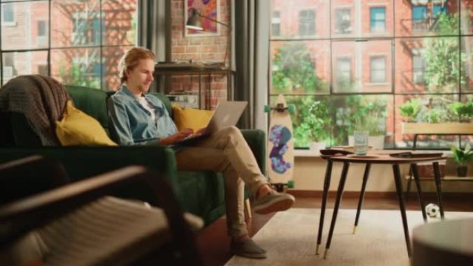 年轻英俊的成年男子坐在阁楼公寓的沙发上，在家用笔记本电脑工作。创意男性查看社交媒体，浏览互联网。从大
