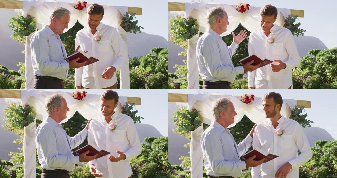 微笑的高加索高级男性婚礼主持人手持书籍和新郎站在户外祭坛上