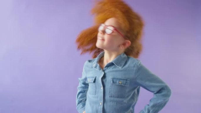 红色头发和眼镜的微笑女孩在紫色工作室背景下摇晃头发的工作室镜头-慢动作拍摄