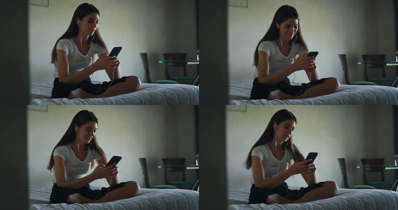 一个年轻女子的真实照片坐在床上，用智能手机在家里的卧室里用耳机听音乐。