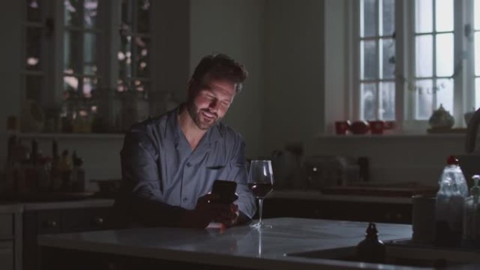 穿着睡衣的成熟男人晚上用手机坐在厨房喝杯酒