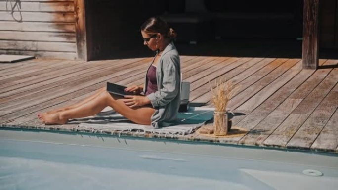 WS年轻女子挂断电话，在游泳池旁的木制露台上打开笔记本电脑