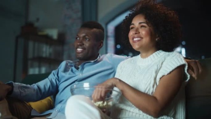 黑人夫妇在电视上看喜剧电影，深夜坐在公寓的沙发上吃爆米花。笑男女朋友在家一起欣赏搞笑电视剧。低角度射