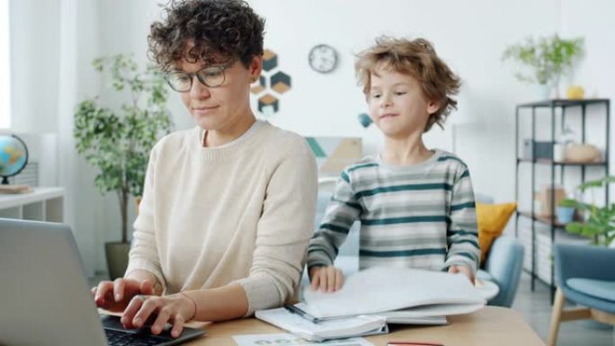 工作的母亲使用笔记本电脑，然后拥抱孩子在公寓里一起画画
