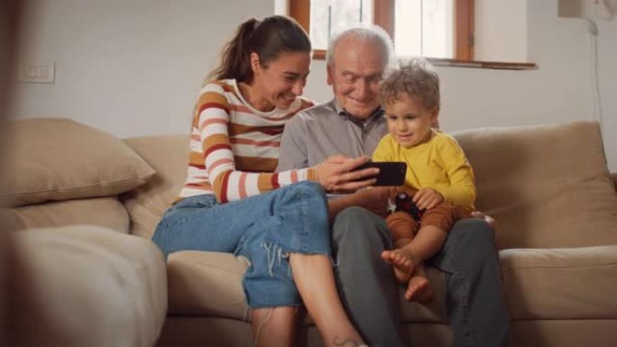 快乐的小家庭聚集在一起，坐在客厅的沙发上，使用智能手机。母亲，儿子和祖父在手机上观看视频并大笑。他们