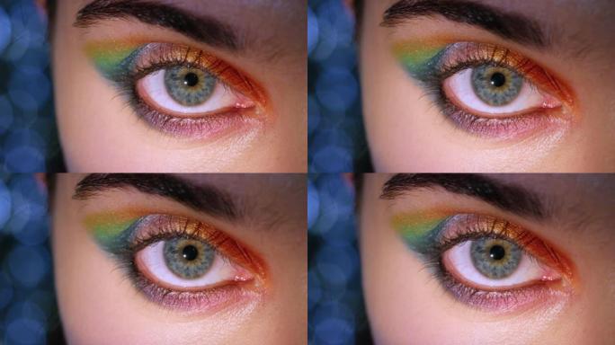 彩虹节化妆的女性眼睛，眼睑上有强烈的鲜艳色彩，细节为蓝绿色的瞳孔和虹膜。特写镜头显示眉毛上的微光，健