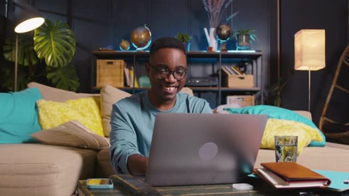 在舒适的家庭房间使用笔记本电脑编写代码，在眼镜上在线工作年轻快乐聪明的黑人自由商人。