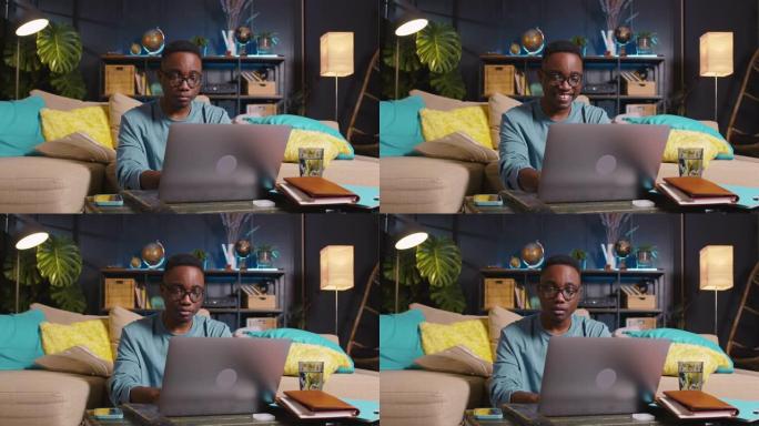 在舒适的家庭房间使用笔记本电脑编写代码，在眼镜上在线工作年轻快乐聪明的黑人自由商人。