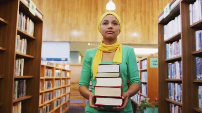 一名戴着黄色头巾的亚洲女学生在书架之间行走，抱着一堆嘘声