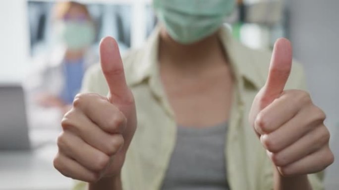 自信的亚洲女医生戴着防护口罩，年轻的病人女孩在诊所或医院的办公桌上做着竖起大拇指微笑。