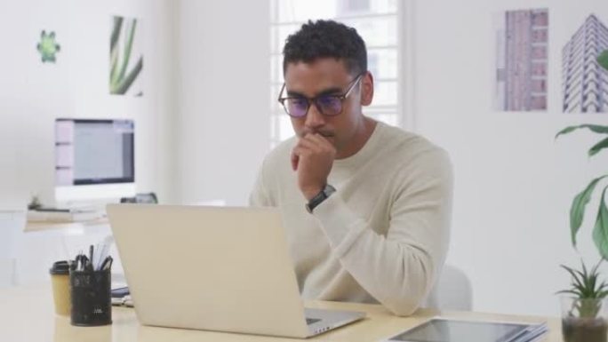 一位专注的商人在办公室的笔记本电脑上工作时会思考想法。年轻的设计师戴着眼镜浏览互联网以获取灵感，同时