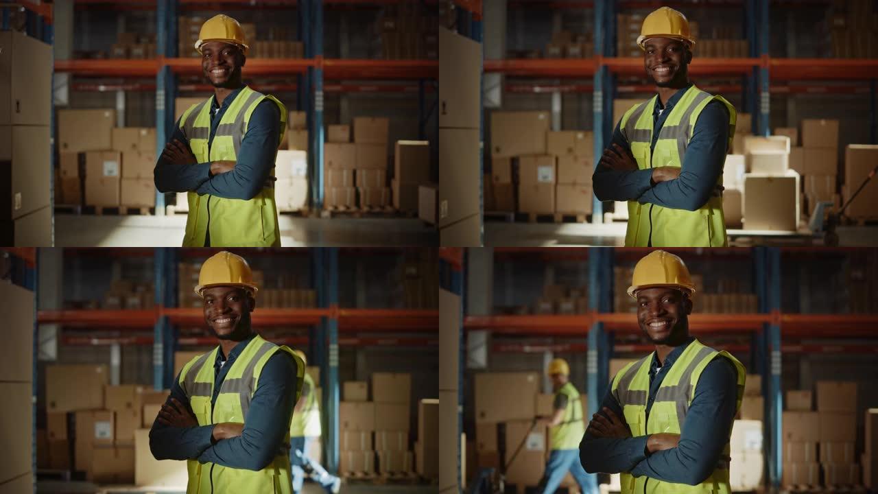 英俊快乐的职业工人穿着安全背心，戴着安全帽，微笑着在镜头前交叉双臂。在后台的大仓库里，货架上堆满了送