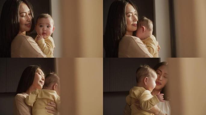 一位美丽的年轻亚洲妇女站在家里的窗户旁边时，抱着婴儿的肖像。可爱的小孩在母亲的怀抱中休息，看着相机