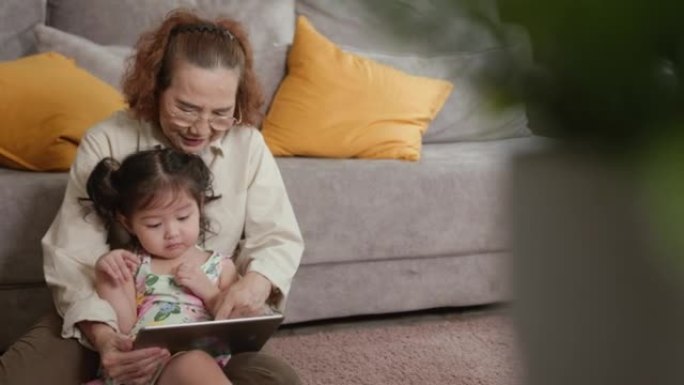 快乐的奶奶在家和可爱的孙女玩数字平板电脑。老年人身心锻炼的机会