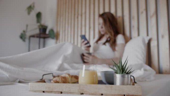 早餐托盘在床上，女人在背景中使用手机和喝咖啡