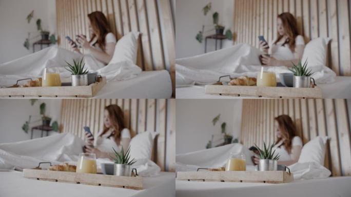 早餐托盘在床上，女人在背景中使用手机和喝咖啡