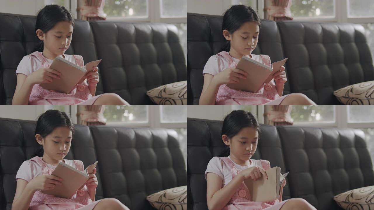 亚洲小女孩坐在沙发上看书，呆在家里。她翻开学习书的一页。学习教育书籍的女孩。