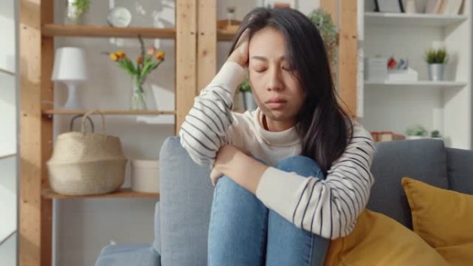 体贴的亚洲女士坐在客厅沙发上拥抱膝盖，看着外面，感到孤独，悲伤沮丧的少年独自呆在家里。