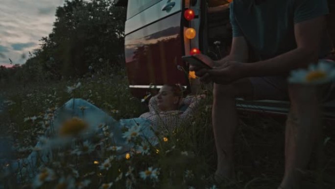 当女人躺在黄昏时停在草地上的露营者旁边时，DS男人使用智能手机