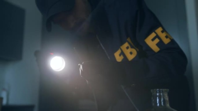 特写一名FBI探员穿着制服，戴着帽子，拿着手电筒，在一个黑暗的房间里，他戴着手套，手里拿着一个弹壳