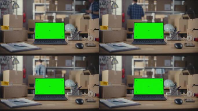 笔记本电脑站在一张绿屏Chromakey模拟显示器的桌子上。小型企业仓库，工人在后台行走。带纸箱的桌